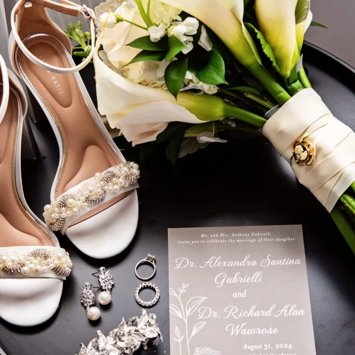 Flat lay of elegant bridal accessories including white pearl-embellished heels, a calla lily bouquet, wedding jewelry, and a wedding invitation for Dr. Alexandra Gabrielli and Dr. Richard Wawrose dated August 31, 2024.