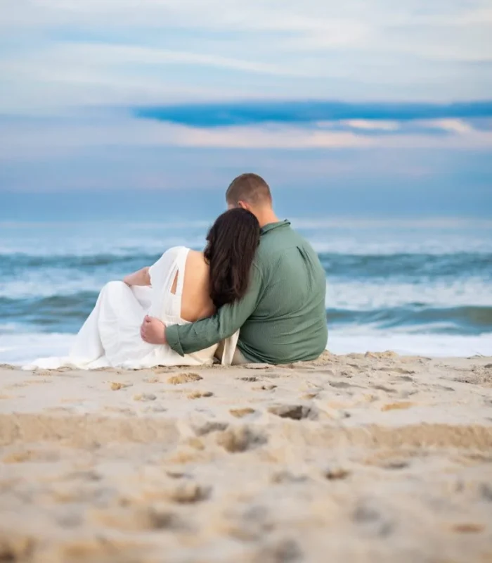 Romantic couple sitting close together on a sandy beach, facing the ocean at sunset with peaceful waves in the background.