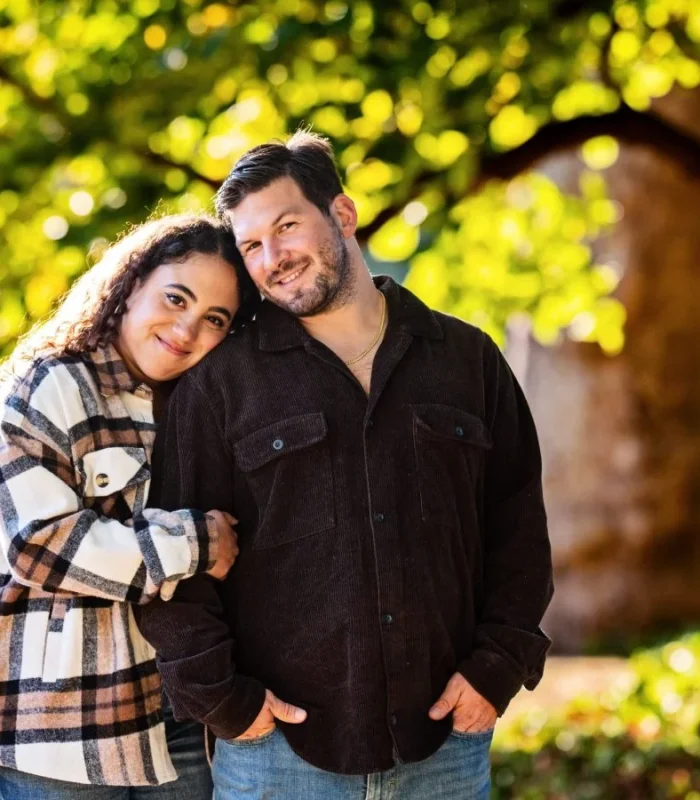 Happy couple posing outdoors in a sunny park, with the woman leaning her head affectionately on the man's shoulder.
