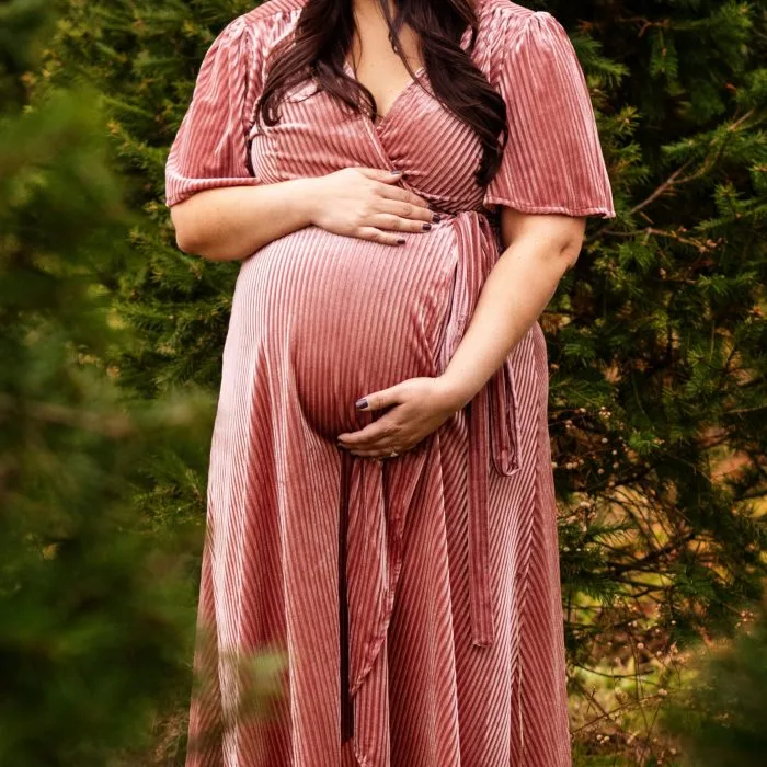 Pregnant woman in a pink striped dress gently holding her baby bump while standing outdoors among green trees during her maternity photoshoot.