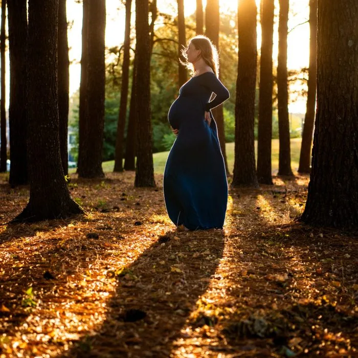 Pregnant woman in a long dark blue dress standing in a sunlit forest, gently holding her baby bump as golden sunlight filters through the trees.