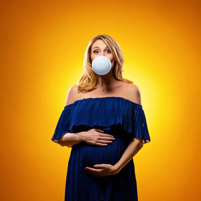 Playful pregnant woman in an off-shoulder blue dress blowing a bubble with gum, standing against a vibrant orange background.