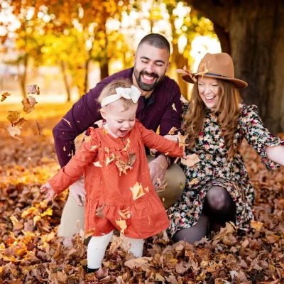 Joyful family playing in autumn leaves during its family photography — smiling mother, father, and young daughter enjoying fall together under a large tree