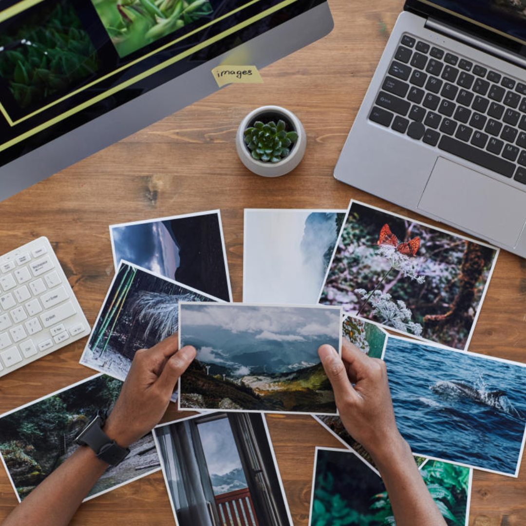 Photographer or designer selecting and organizing printed photos on a wooden desk next to a laptop.
