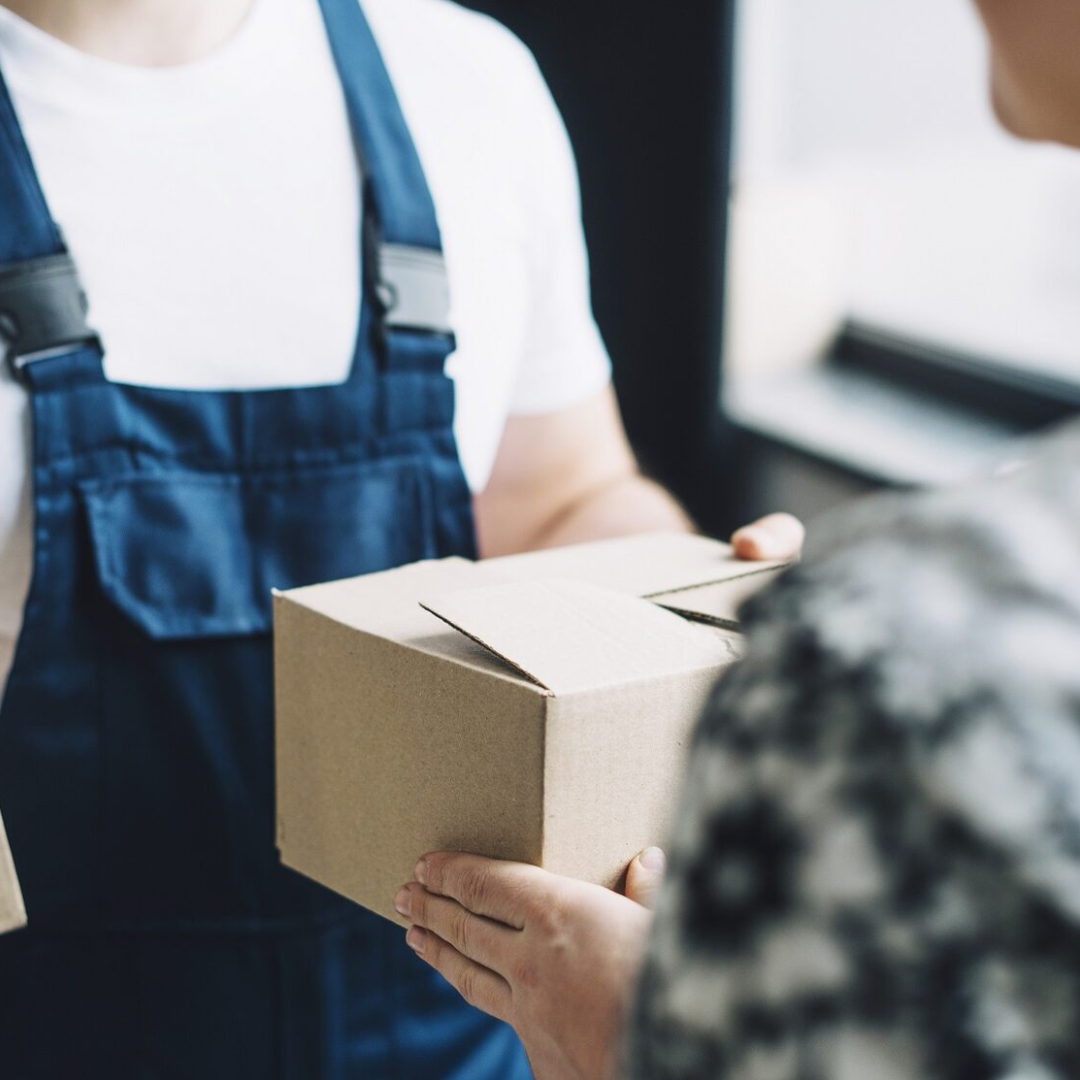 A photo delivery person in blue overalls hands a cardboard package to a customer.