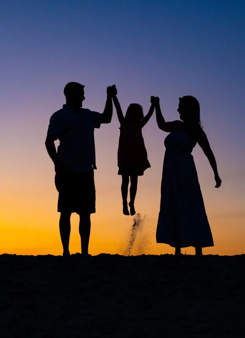 Heartwarming family photography of parents and child enjoying a beach sunset, with the child playfully jumping while holding their hands.