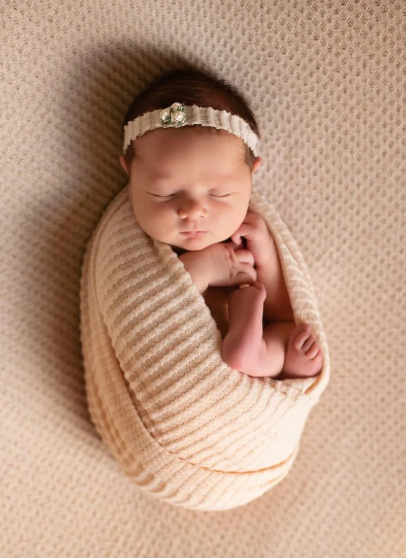 Newborn photography of a peacefully sleeping baby wrapped in a soft, beige swaddle with a delicate headband, lying on a textured blanket.