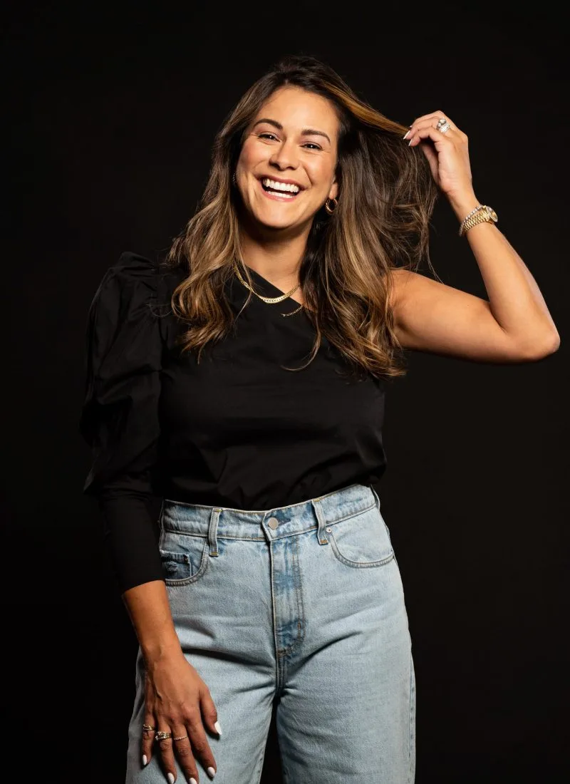 Portrait photography of a smiling woman in a black top and jeans, playfully touching her hair against a dark background.